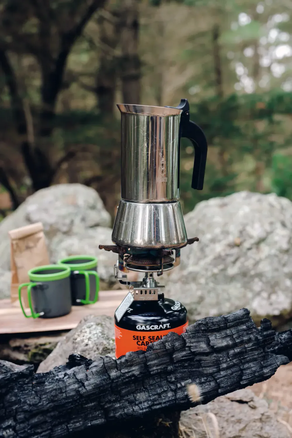 A portable stove with a metallic coffee pot on top, set on a charred log in a forested outdoor area, with coffee mugs and a paper bag in the background.