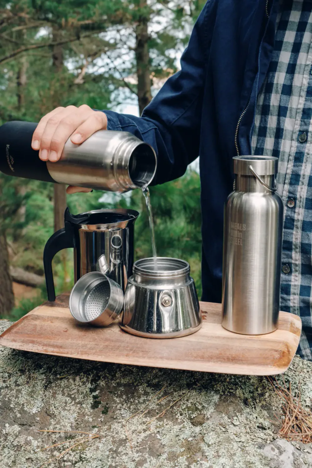 Person pouring water from a thermos into a coffee dripper on a wooden table outdoors with trees in the background.