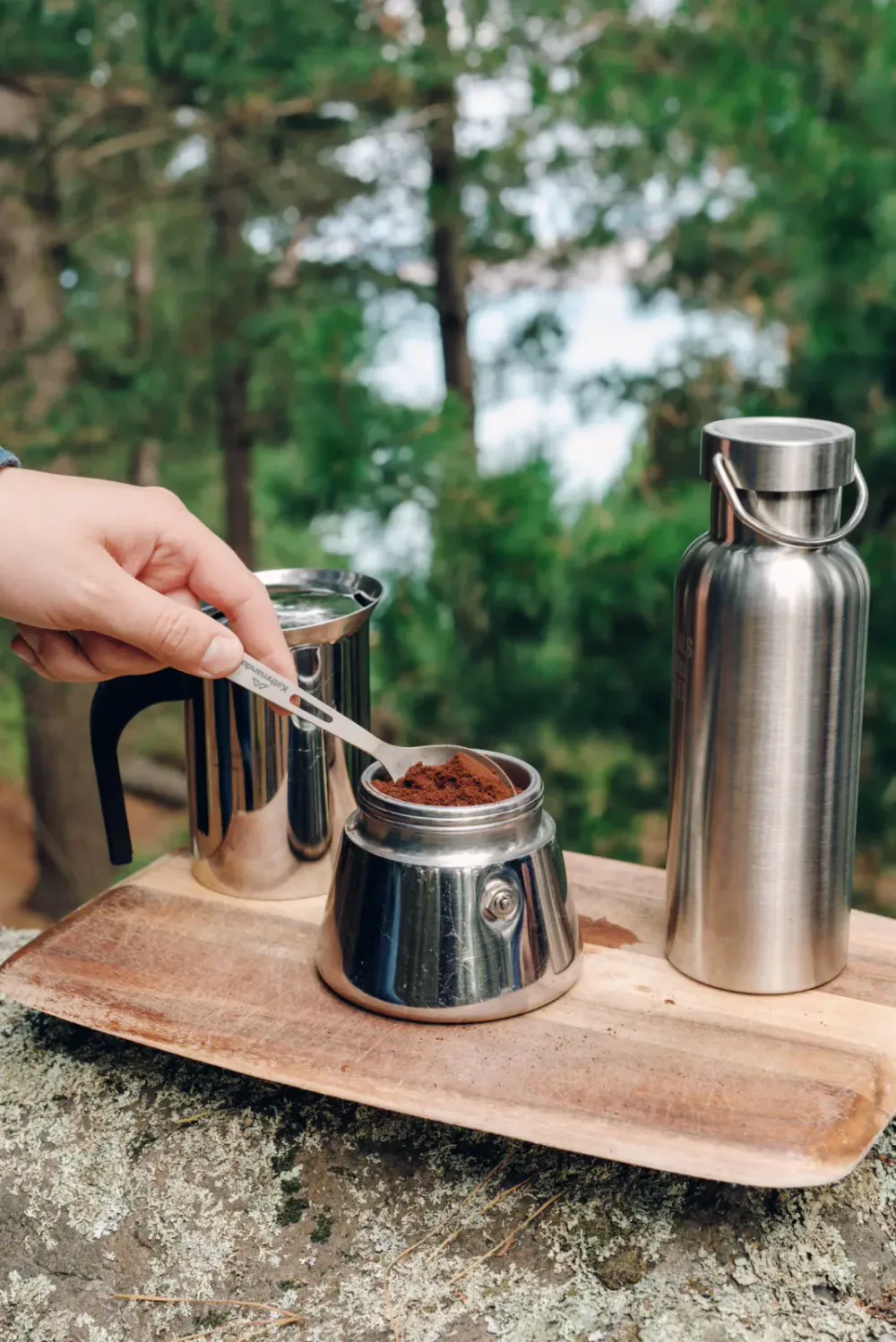 Person scooping coffee grounds from a jar onto a metal measuring spoon, with a thermos and stainless steel water bottle on a wooden tray outdoors in a forest.