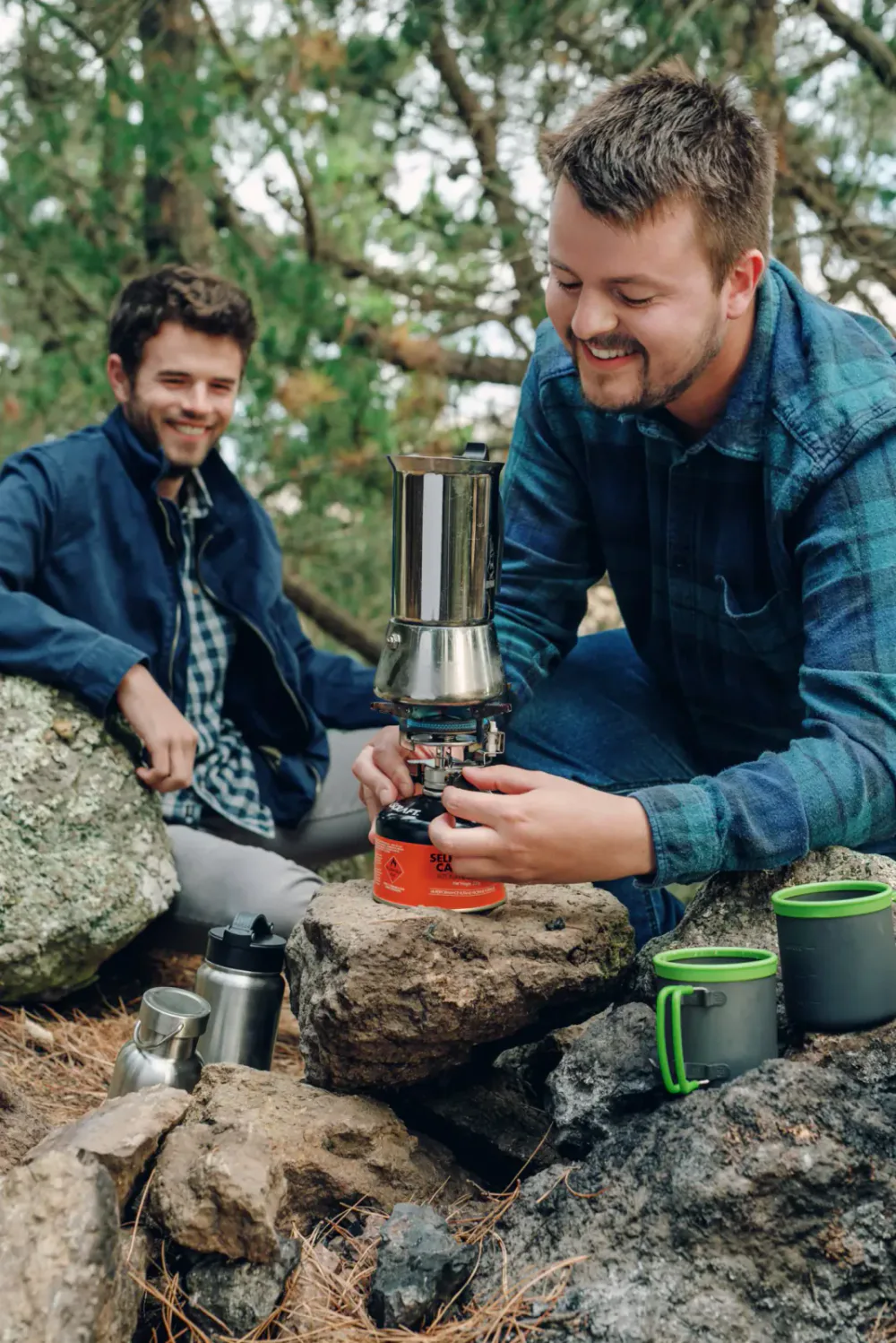 Two men camping outdoors surrounded by trees; one is preparing a coffee or hot meal using a portable stove on a rock, while the other watches and smiles.