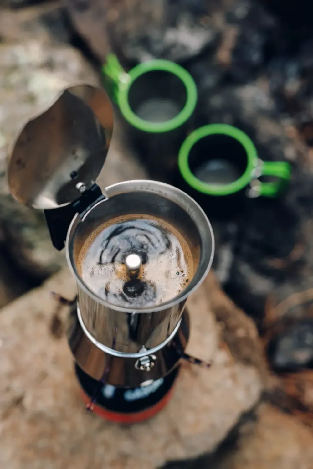 A close-up of a metal camping kettle boiling coffee over a small portable stove, with two green camping mugs in the background outdoors.