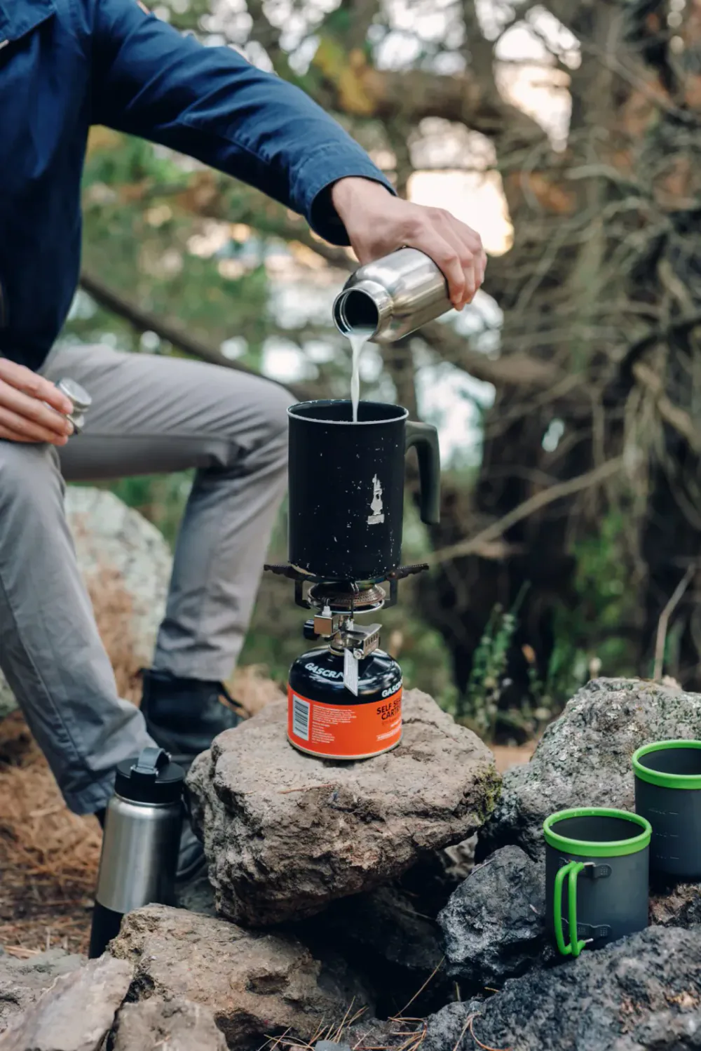Person pouring liquid from a thermos into a mug on a portable camping stove outdoors surrounded by rocks and trees.
