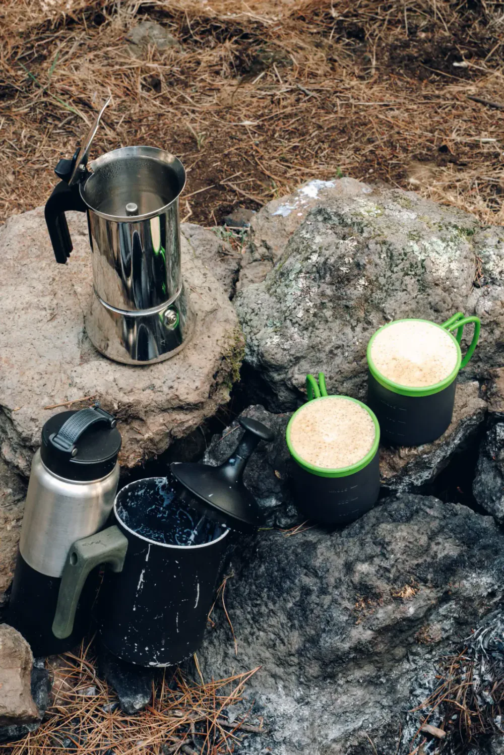 Camping coffee setup with a moka pot, two mugs filled with coffee, and a thermos on rocks surrounded by pine needles.
