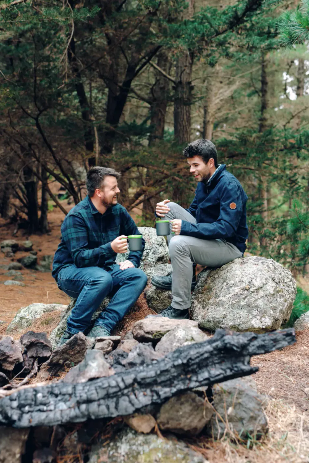 Two men sitting on rocks in a forest, smiling and talking while holding mugs.