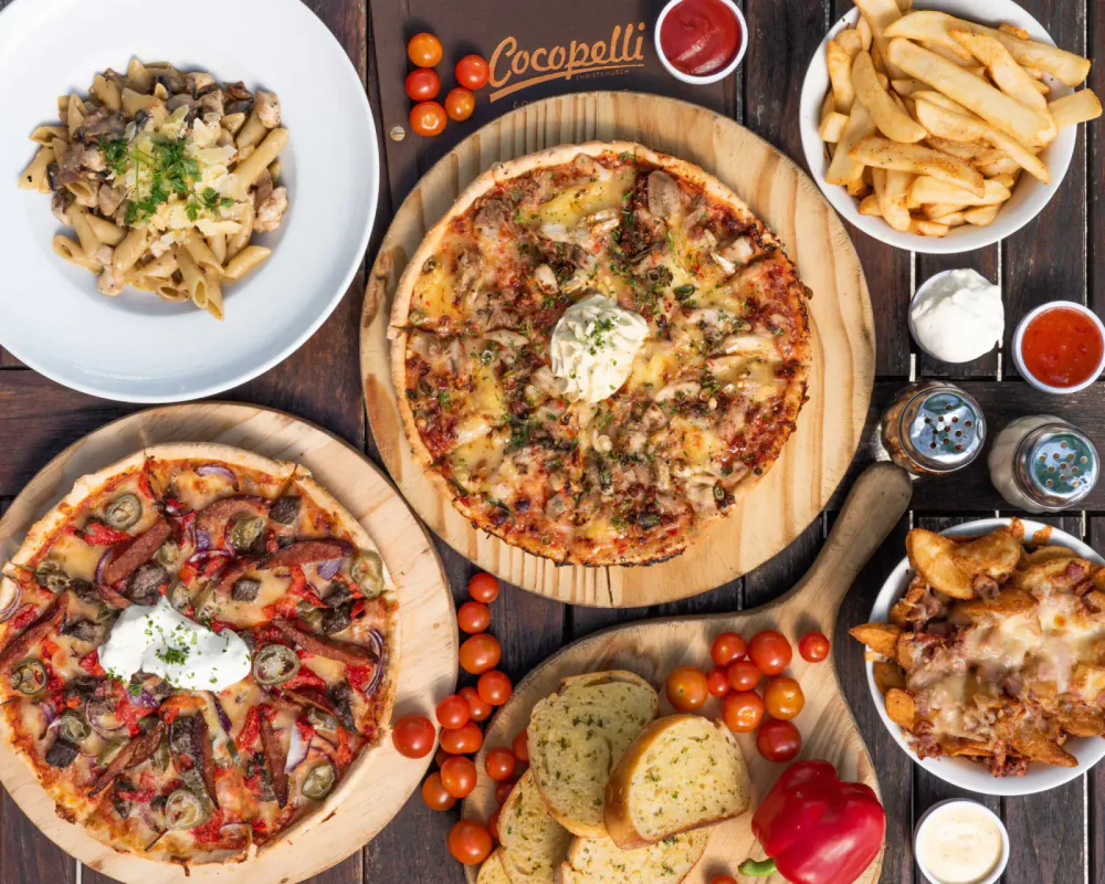 A variety of Italian dishes including two pizzas, a pasta dish, French fries, garlic bread, cherry tomatoes, a red bell pepper, and small containers of sauces and dips on a dark wood table.