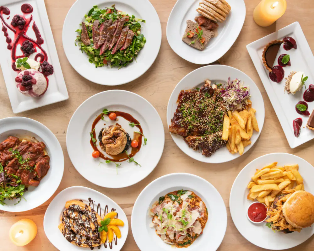 A variety of dishes on white plates, including salads, meat dishes, pasta, fries, dessert, and a burger, arranged on a wooden table with candles.