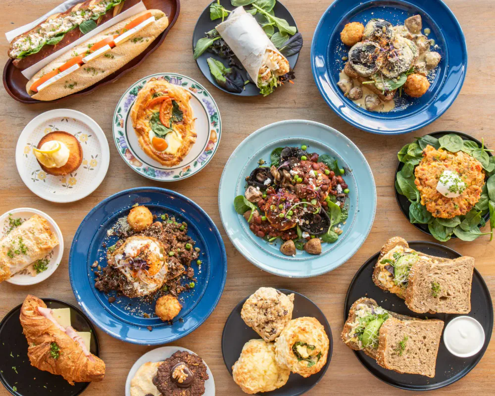 A variety of breakfast and brunch dishes on a wooden table, including sandwiches, salads, a pastry, a cupcake, a croissant, and other baked goods.