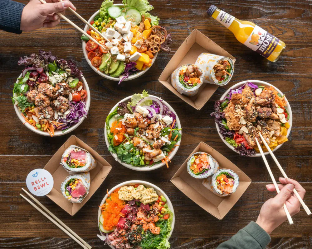A variety of colorful bowls of Asian cuisine and sushi wraps on a wooden table, with people using chopsticks.