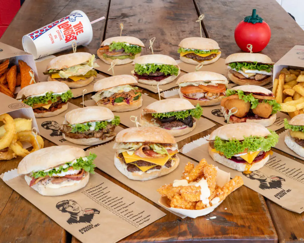 Variety of burgers, fries, onion rings, and chicken tenders on a wooden table with a paper menu and a drink.