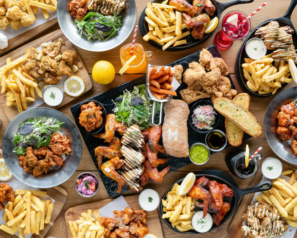 An overhead view of a variety of dishes including fried chicken, fries, salads, bread, and beverages on a wooden table at a restaurant or gathering.