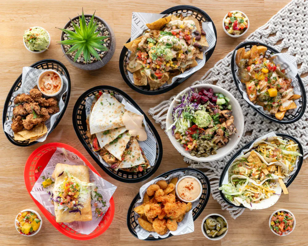 A variety of Mexican dishes on a wooden table, including tacos, nachos, fried chicken, tortilla wrap, salad, and bowls of salsa and guacamole.