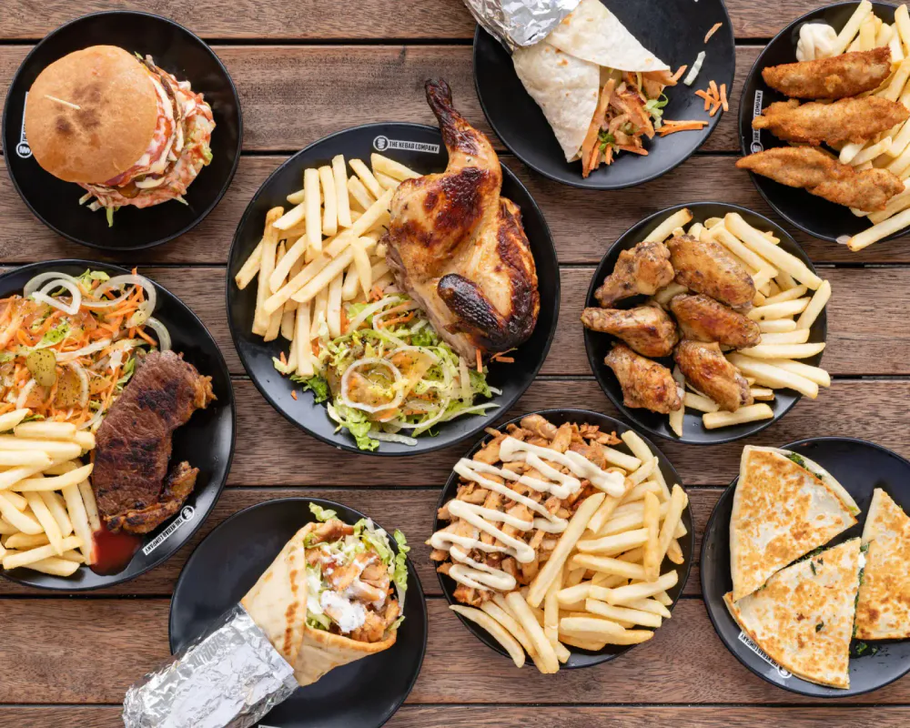 Assorted plates of fried chicken wings, fries, grilled chicken, a burger, a wrap, quesadilla, and salad on a wooden table.