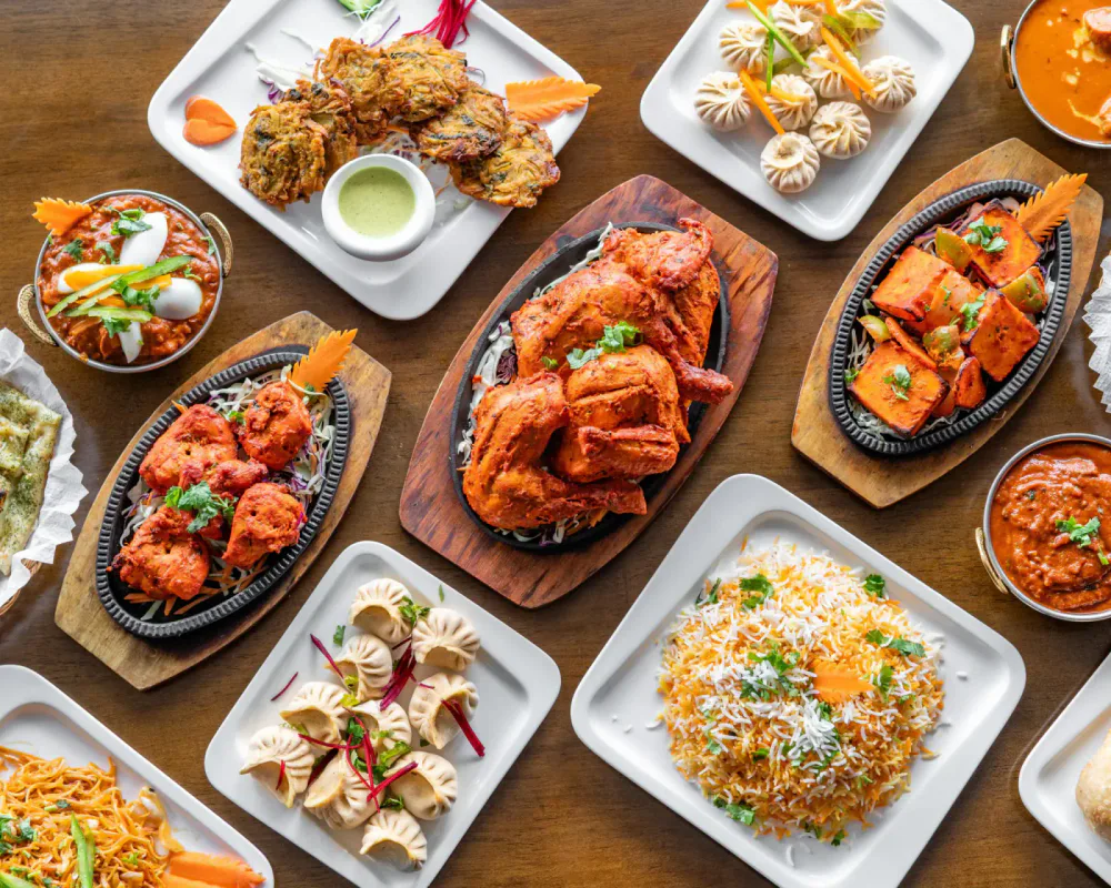 A variety of Indian dishes on white and black plates, including tandoori chicken, curries, samosas, naan bread, and rice, all arranged on a wooden table.