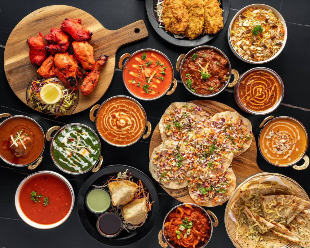 Assorted Indian dishes including chicken tikka, fried chicken, vegetable biryani, various curries, naan bread, samosas, and sauces on a black surface.