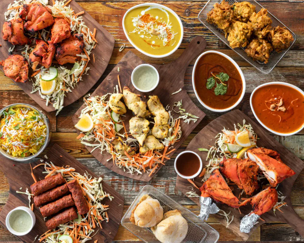 An Indian feast with various dishes including chicken tikka, samosas, dal, curries, rice, and grilled chicken served on wooden platters and bowls.