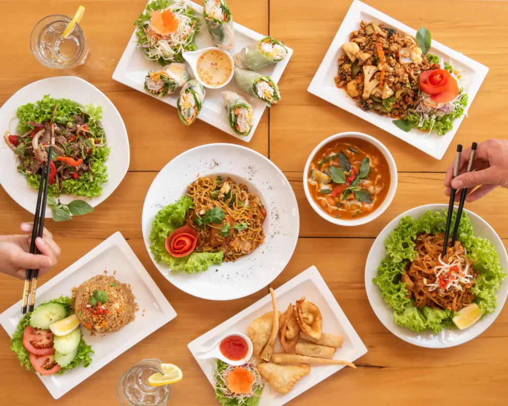 Assorted Asian dishes including spring rolls, stir-fried noodles, salad, curry, and fried items on a wooden table, with glasses of water.