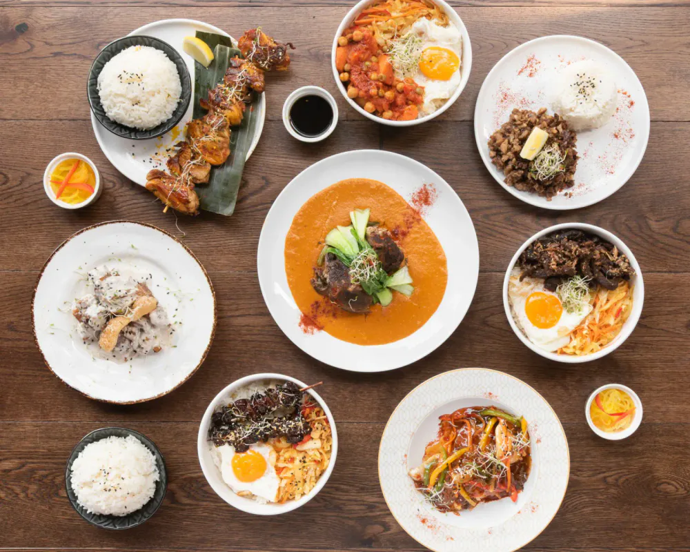 Assorted Indian dishes on a wooden table including rice, chicken skewers, curry, beef and vegetable dishes with noodles, and small bowls of pickles and sauces.