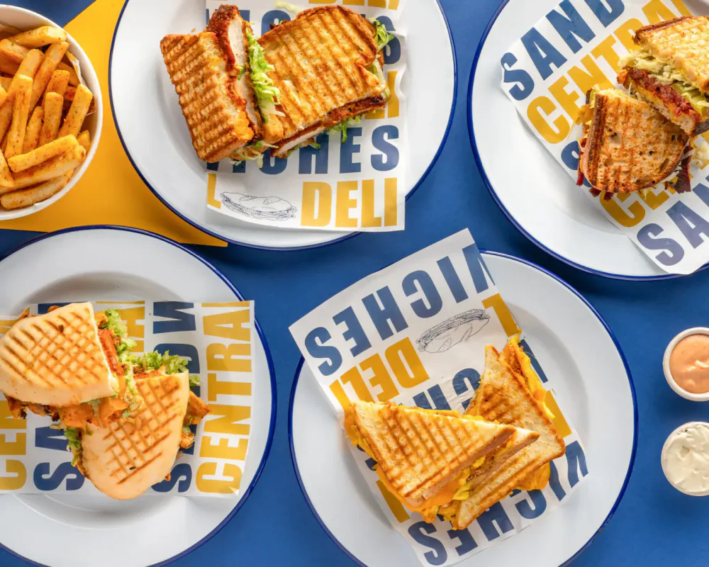 Four freshly prepared sandwiches served with condiments and sides on a blue table.