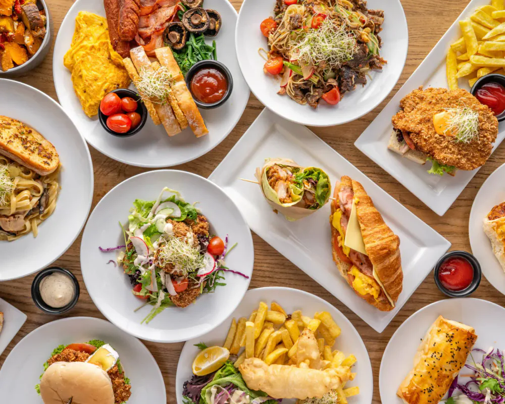 A table filled with various plates of American-style food including salads, sandwiches, fries, fried chicken, pasta, and breakfast items.