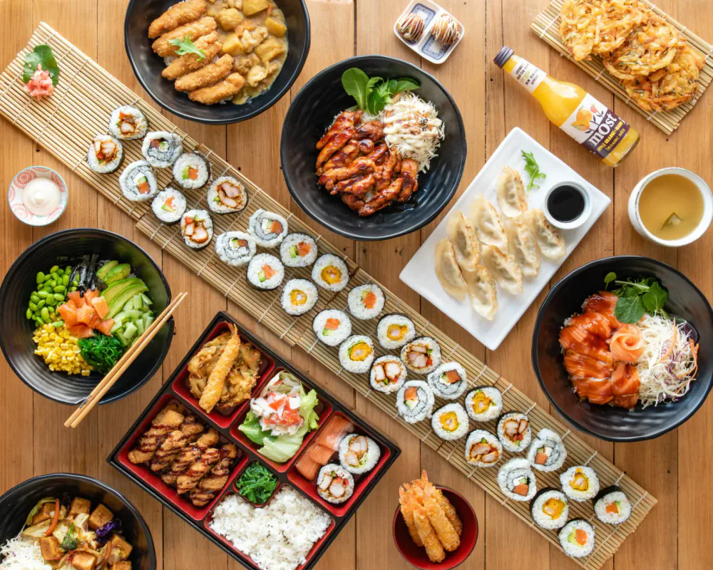 A variety of Japanese dishes on a wooden table, including sushi rolls, sashimi, gyoza, tempura, salad, and drinks.