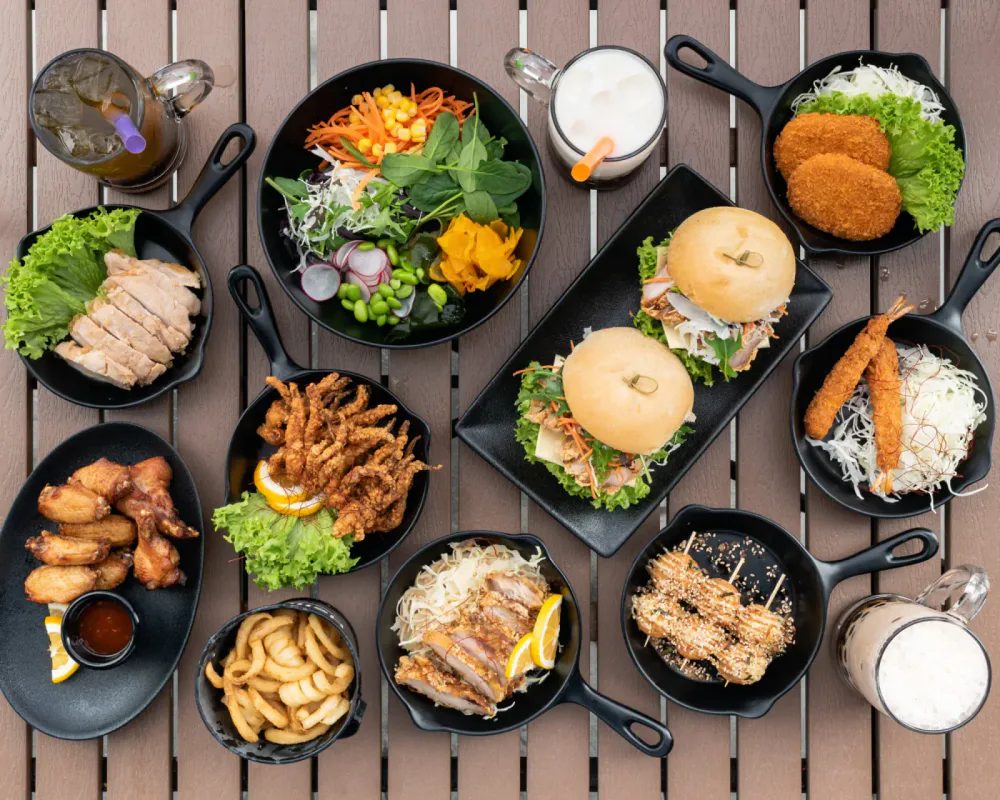 Assorted Asian dishes on a wooden table including fried chicken wings, salad, sandwiches, fried shrimp, onion rings, and beverages.