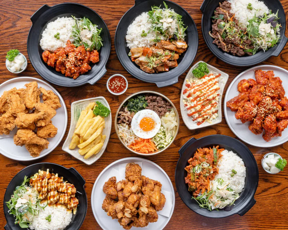 A variety of Korean dishes on a wooden table, including fried chicken, fried potatoes, bibimbap, various stir-fried vegetables, and other Korean-style dishes with rice and sauces.