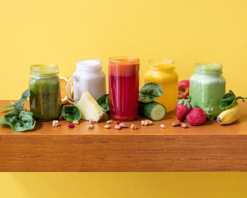 Five jars of colorful smoothies with various fruits and vegetables on a wooden table against a yellow background.