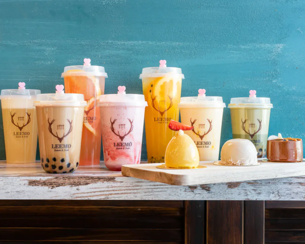 A variety of colorful bubble tea drinks with different flavors and toppings, displayed on a rustic wooden table against a teal wall backdrop, with some desserts on a wooden platter in front.