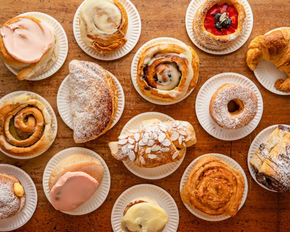 Assorted pastries and sweet baked goods on white paper plates on a wooden surface, including cinnamon rolls, danishes, donuts, croissants, and éclairs.
