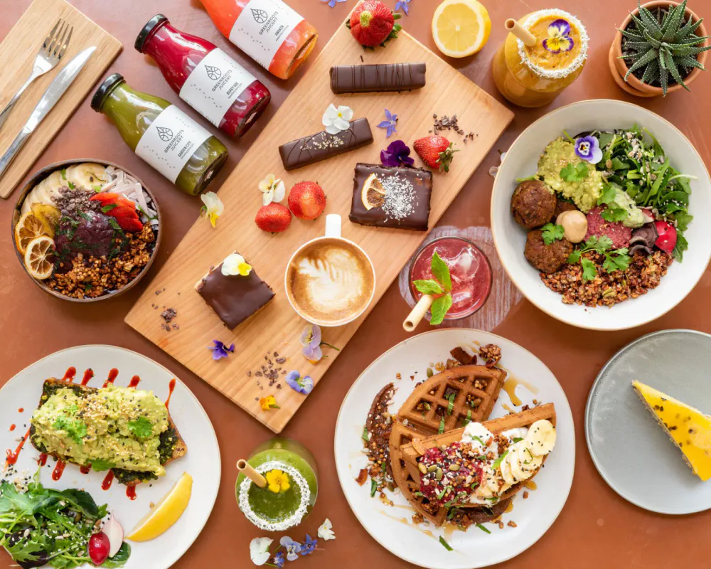 A colorful assortment of healthy foods and desserts on a table, including salads with avocado and vegetables, assorted chocolates and strawberries, a latte, juice bottles, and waffle with banana and berries, decorated with flowers and herbs.