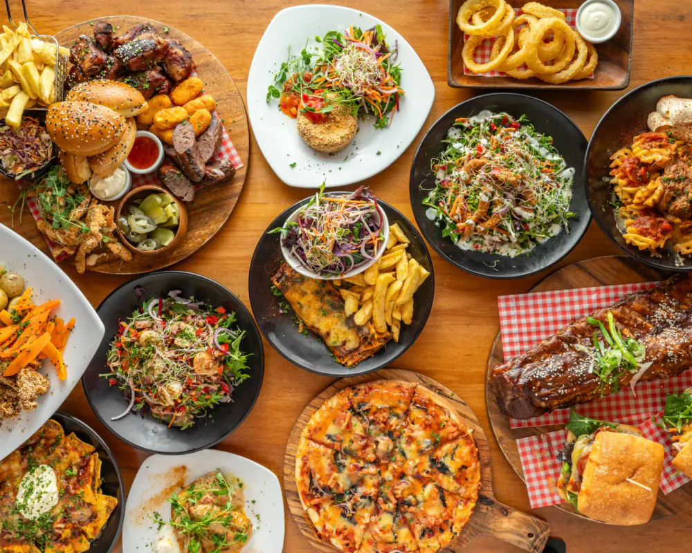 A variety of dishes including pizza, salads, fried chicken, ribs, onion rings, and fries on a wooden table.