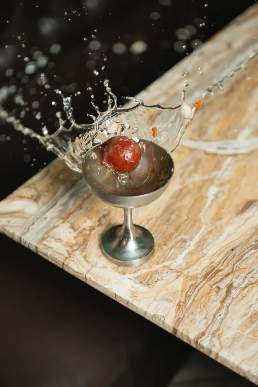 A metallic drink glass on a marble surface with water splashing and a red cherry inside.