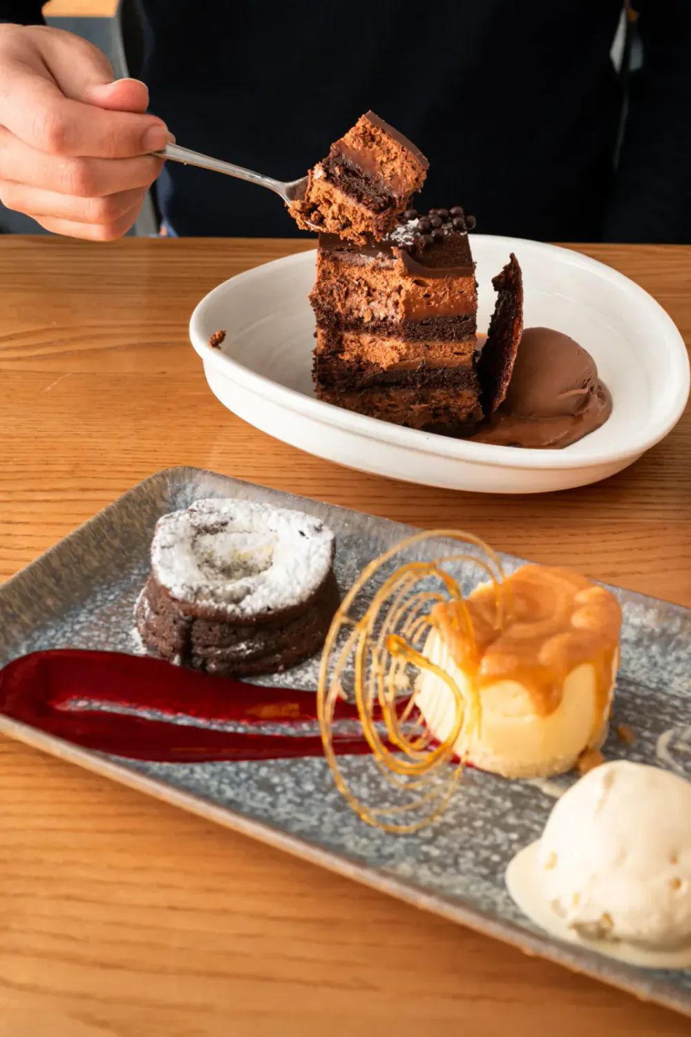 A diner lifts a bite of layered chocolate cake beside a plated chocolate dessert.