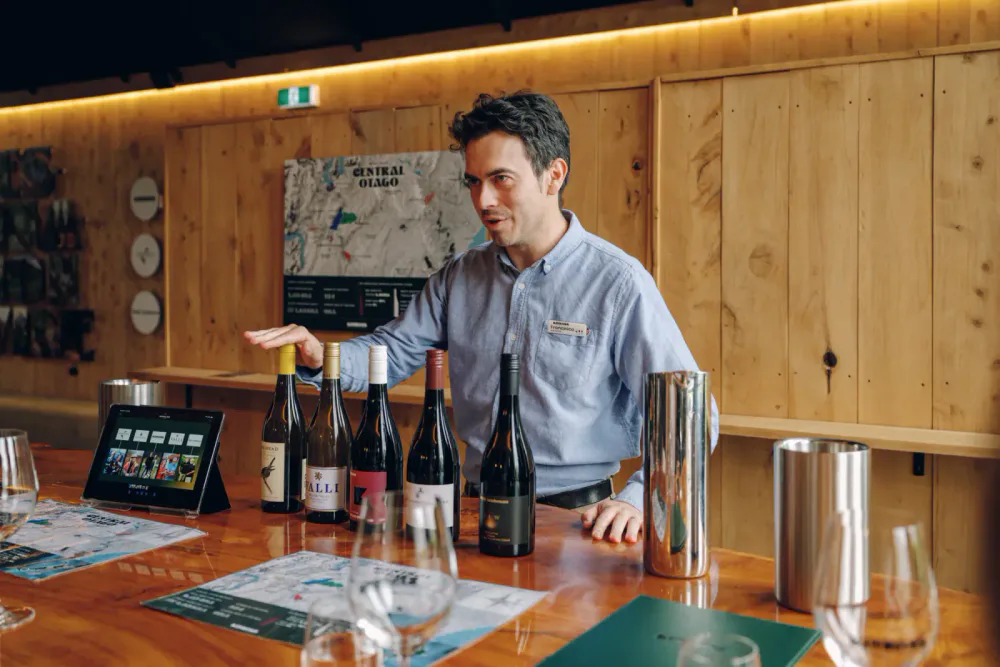 A hospitality staff member standing behind a timber bar with wine bottles, glasses and a point-of-sale tablet