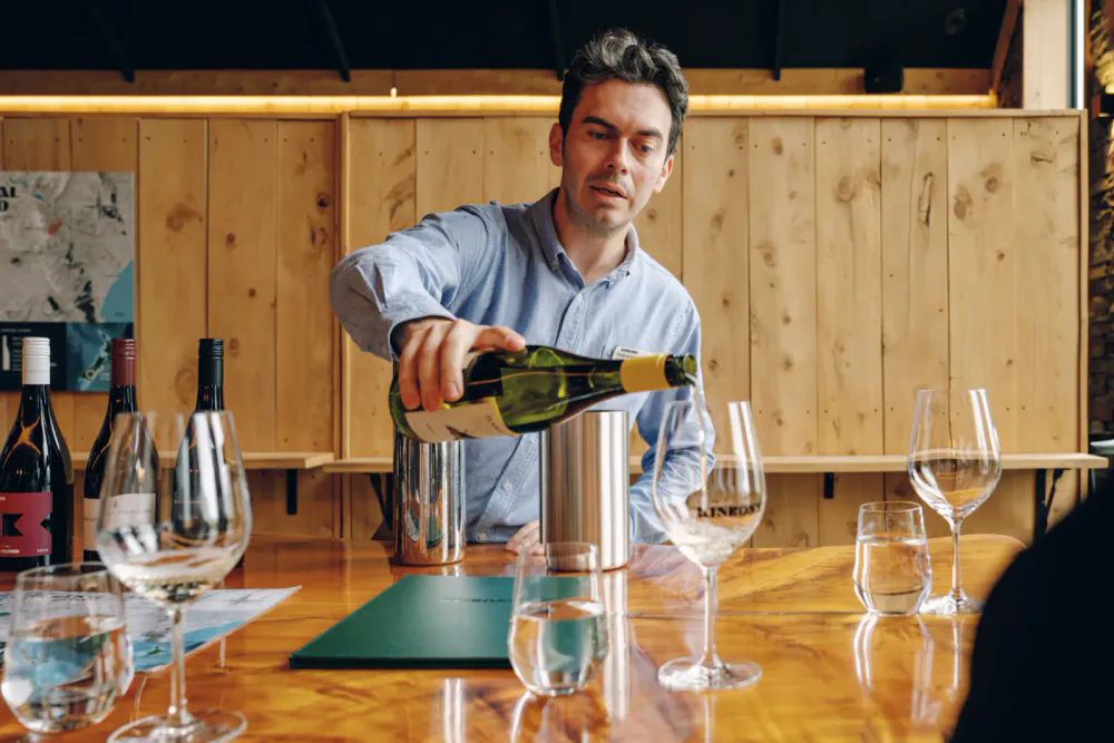 Man pouring white wine into a glass at a wine tasting event with various wine bottles and glasses on the table.