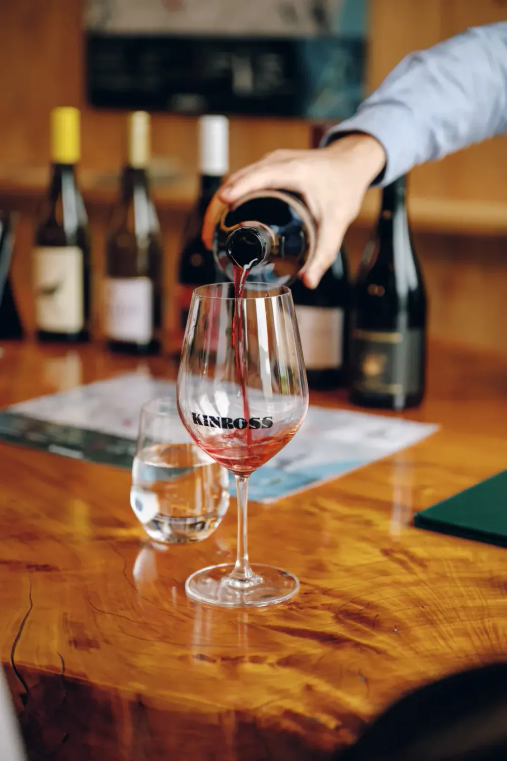 A hand pouring red wine into a wine glass on a wooden table, with several wine bottles in the background.