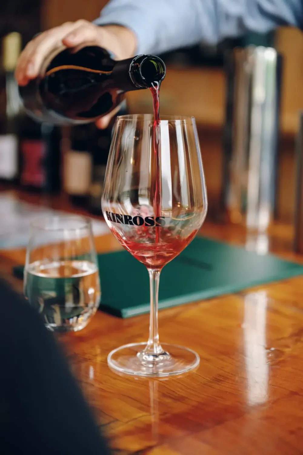 Person pouring red wine into a wine glass on a wooden table, with a glass of water and a green menu nearby.