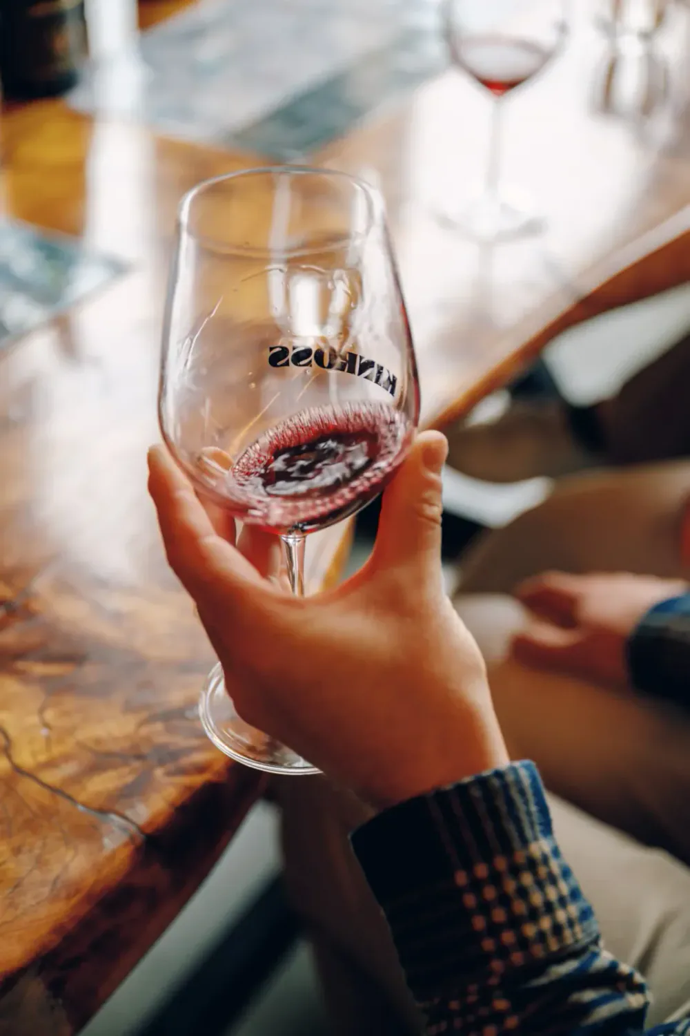 A person holding a wine glass with red wine, with a reflection of the wine in the glass, sitting at a wooden table in a casual setting.