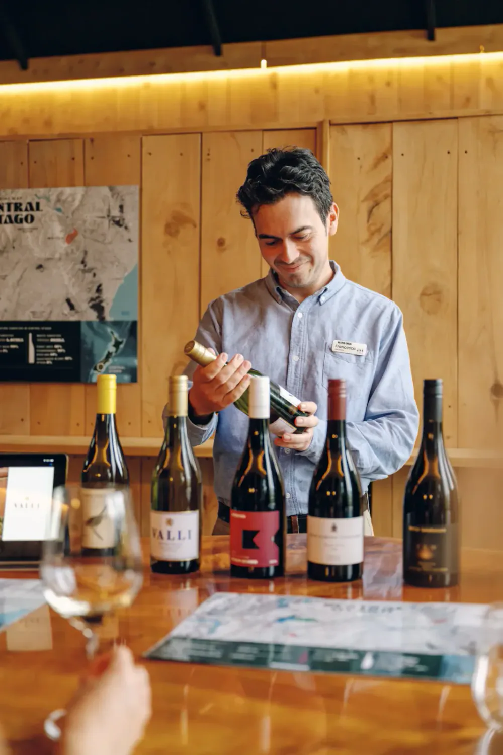 A man with a name tag that reads 'Francesco' standing behind a wooden table, holding a bottle of wine and smiling, with several bottles of wine and glasses on the table.