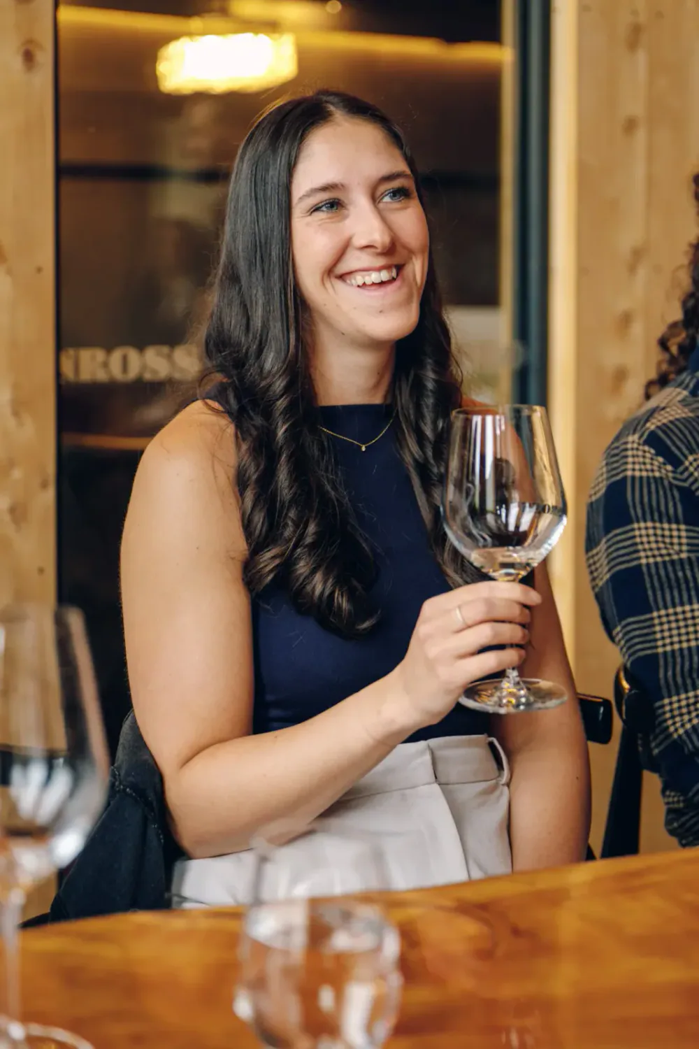 A young woman with long dark hair, wearing a navy sleeveless top and beige pants, sits at a wooden table holding an empty wine glass, smiling in a cozy restaurant with warm lighting.