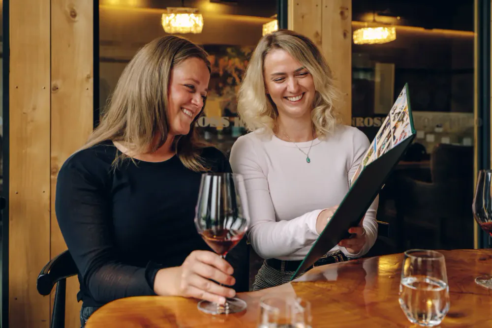 Two women enjoying dinner at a restaurant, looking at a menu and smiling, with glasses of wine on the table.