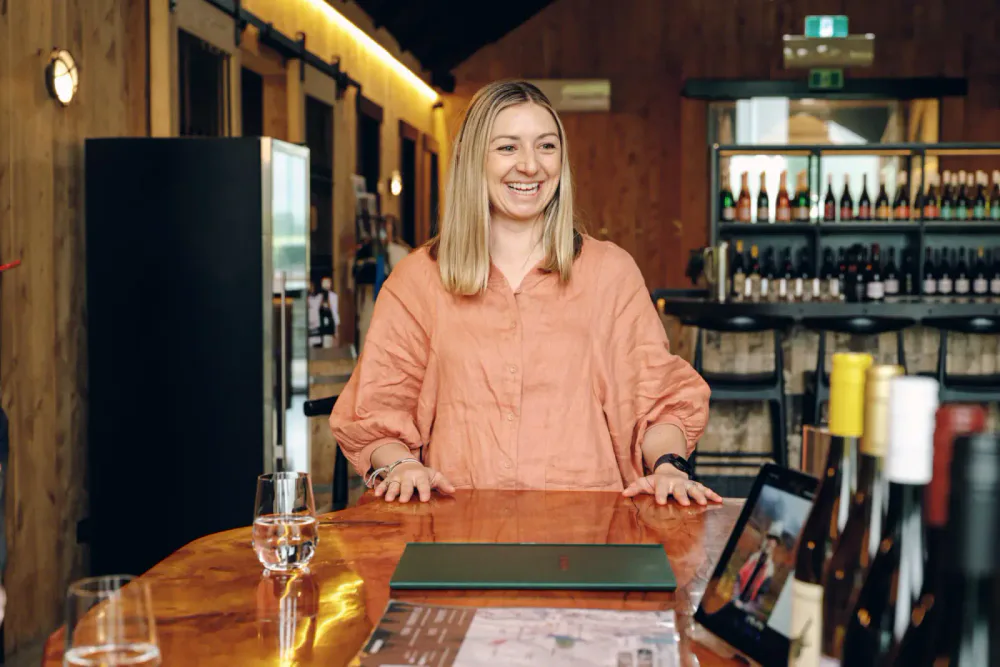 A woman with blonde hair smiling, leaning on a wooden bar counter in a cozy, rustic bar or restaurant. There are glasses and a menu on the counter.