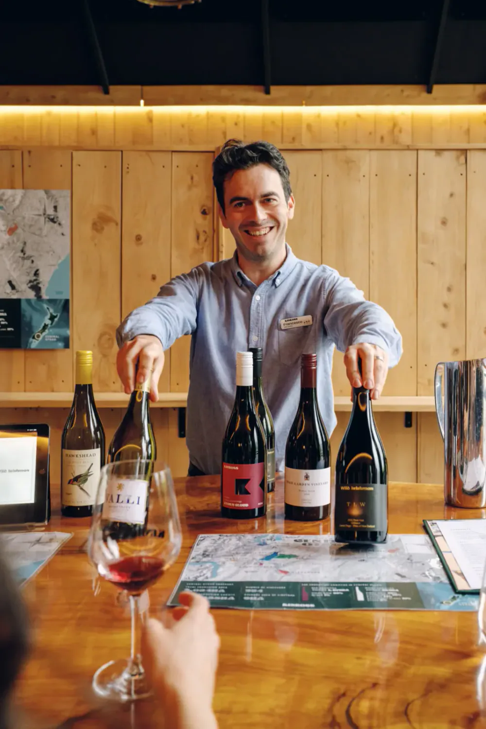 A man with a name tag smiling and standing behind a table with five bottles of wine, offering tastings, in a wooden-walled wine tasting room.
