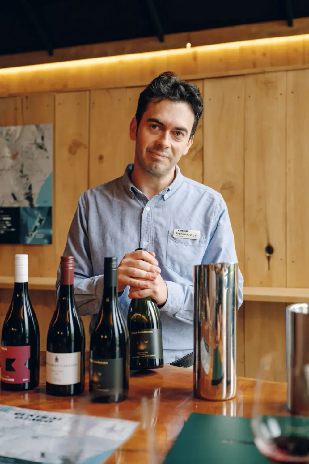 A man wearing a light blue dress shirt with a name tag that says 'Francesco' stands behind a wooden table with several bottles of wine, holding a wine glass and smiling.