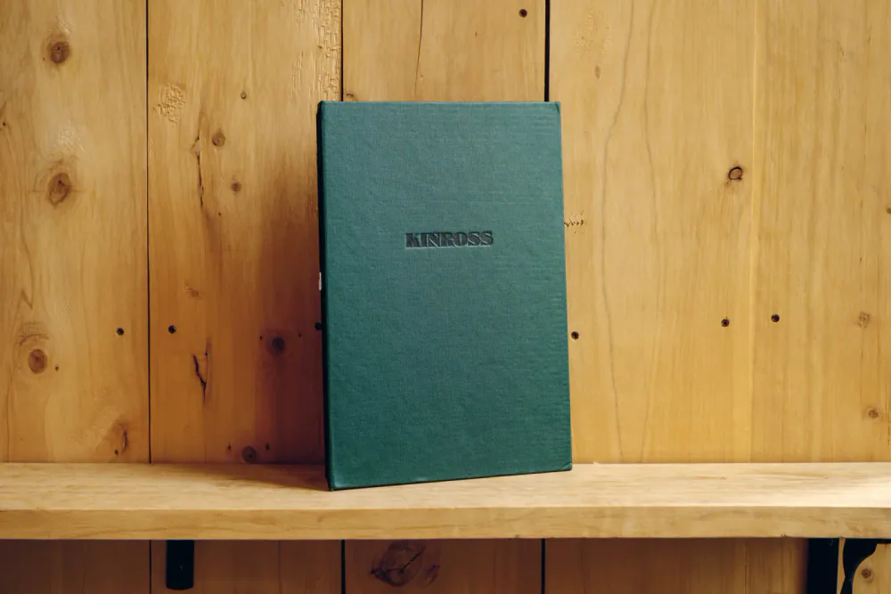 A green box with the word 'KINROSS' on it, standing on a wooden shelf against a wood-paneled wall.