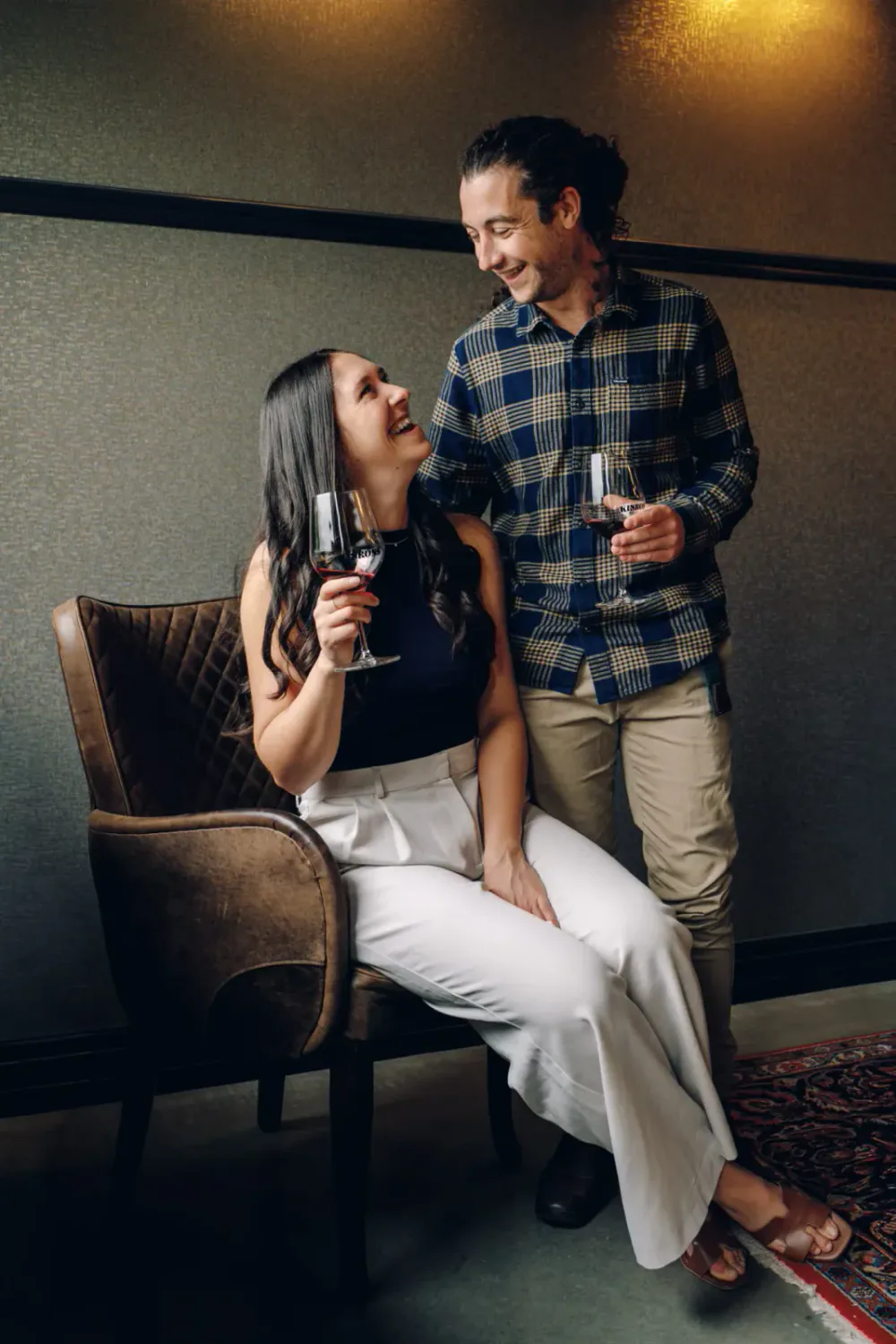 A man and woman sharing a joyful moment with glasses of red wine, sitting on a vintage armchair in a cozy room.