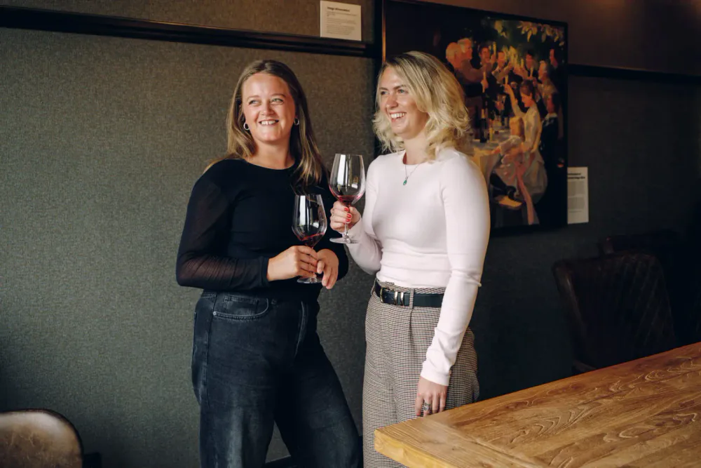 Two women smile over glasses of red wine inside an art-filled hospitality venue.