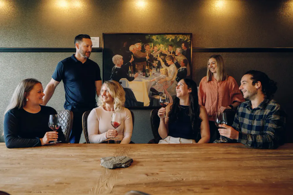 Six people are sitting around a wooden table in a restaurant, holding glasses of red wine and smiling. A man is standing and talking to them. There is a painting of a dinner party hanging on the wall behind them.