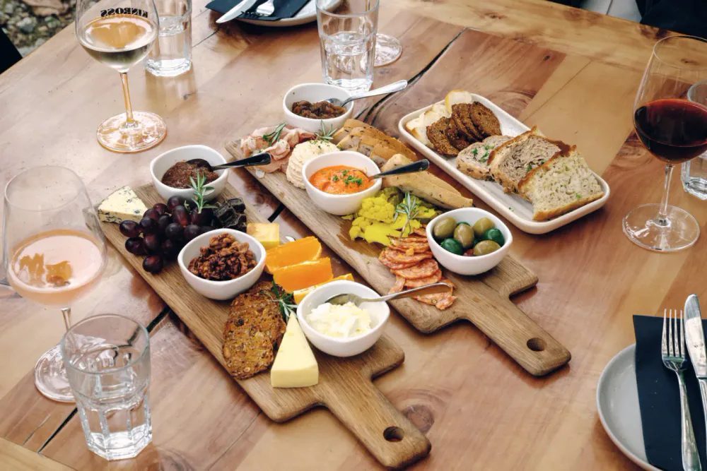 A charcuterie board on a wooden table with cheese, grapes, olives, meats, bread, and dips, surrounded by glasses of white wine, red wine, and water.