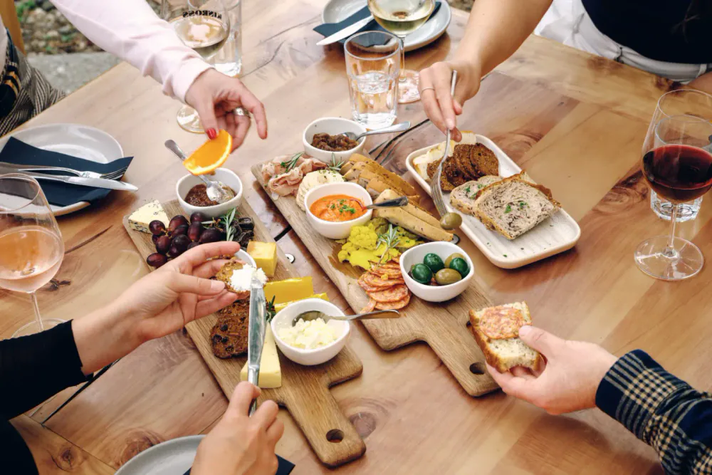 People sharing a cheese and charcuterie board with bread, grapes, olives, spreads, and wine at a wooden table.
